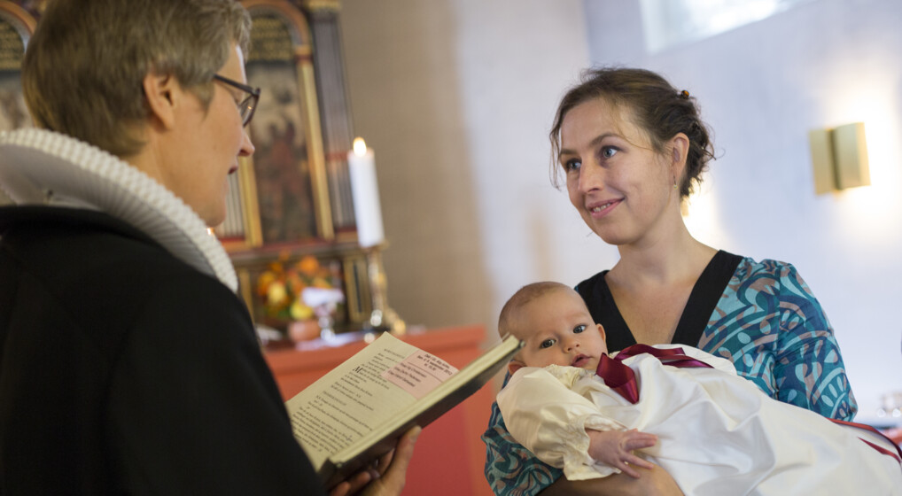 Mor holder dåbsbarn i dåbskjole, mens præst læser fra dåbsritualet