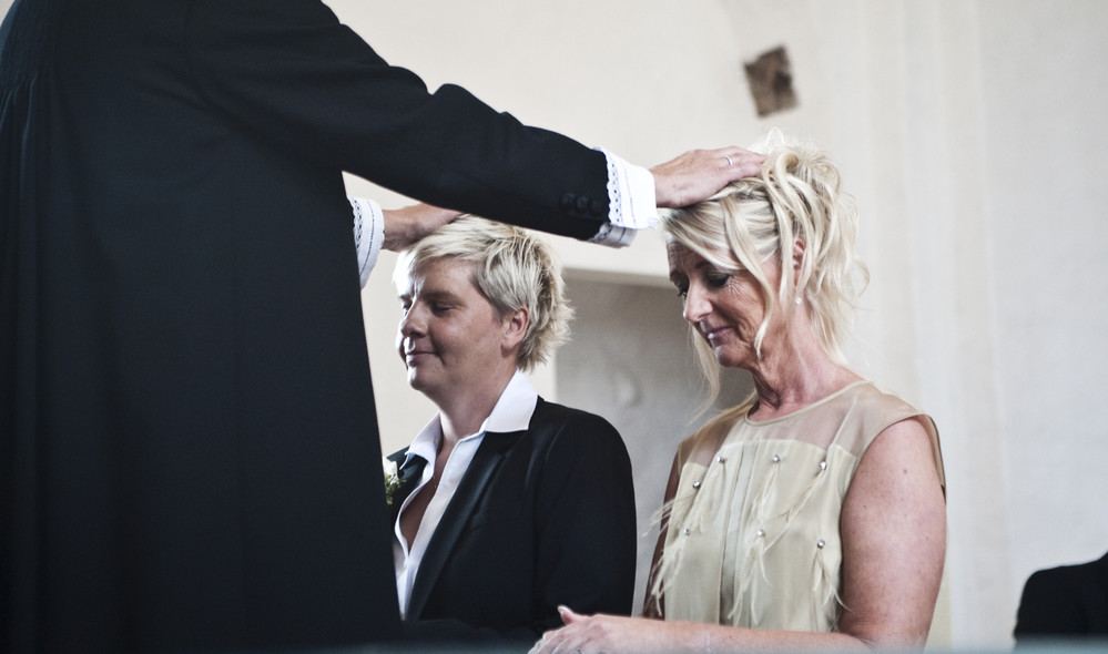 To kvinder knæler foran alteret, præst lægger hænderne på deres hoveder under vielse af par af samme køn i folkekirken